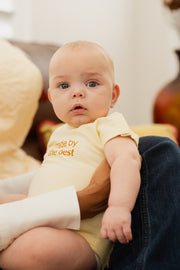 Image of baby with blue eyes wearing cream colored baby onesie with the words "Made by the best" in gold letters